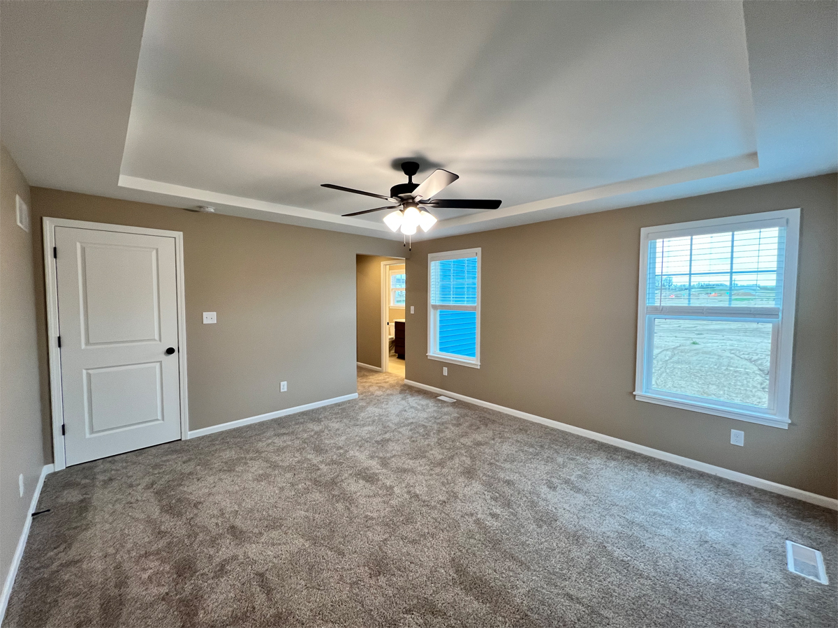 The Redbud master bedroom with carpet, boxed ceiling, ceiling fan, windows and entrance to master bathroom
